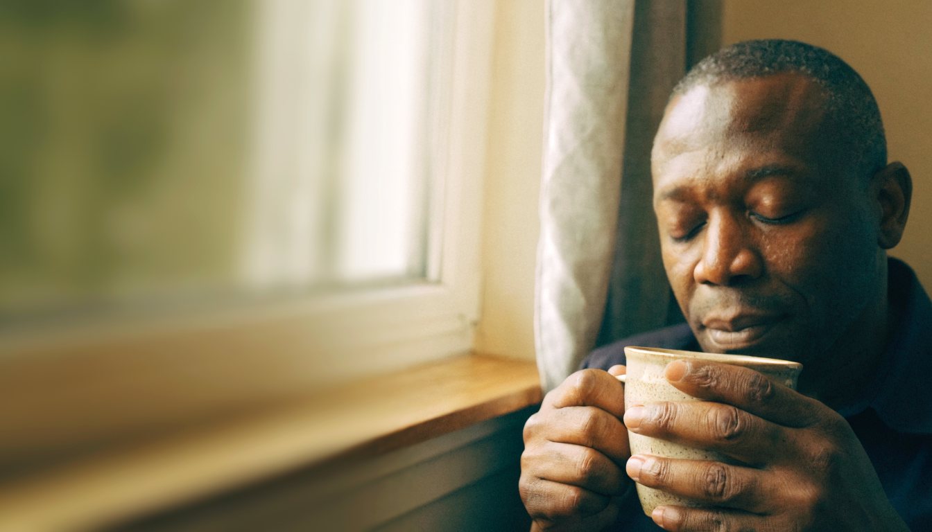 A man sitting quietly by a window at home with a warm drink